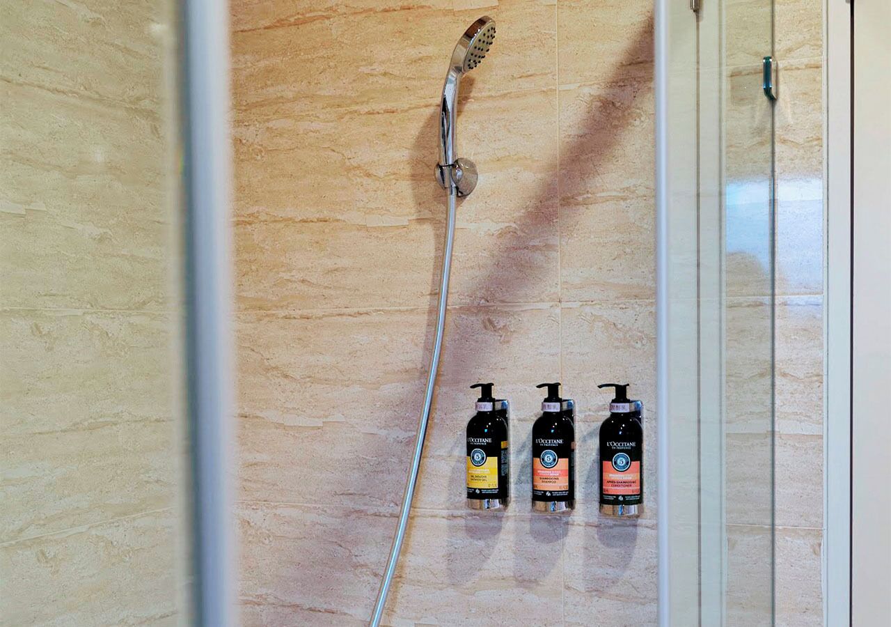 Area doccia con soffione a mano e tre flaconi di bagnoschiuma L'Occitane a parete.