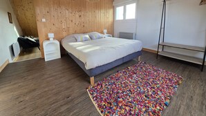 Une chambre avec un plafond en bois, un lit recouvert d’une couette blanche, un tapis coloré, un lampadaire et une fenêtre avec des stores.