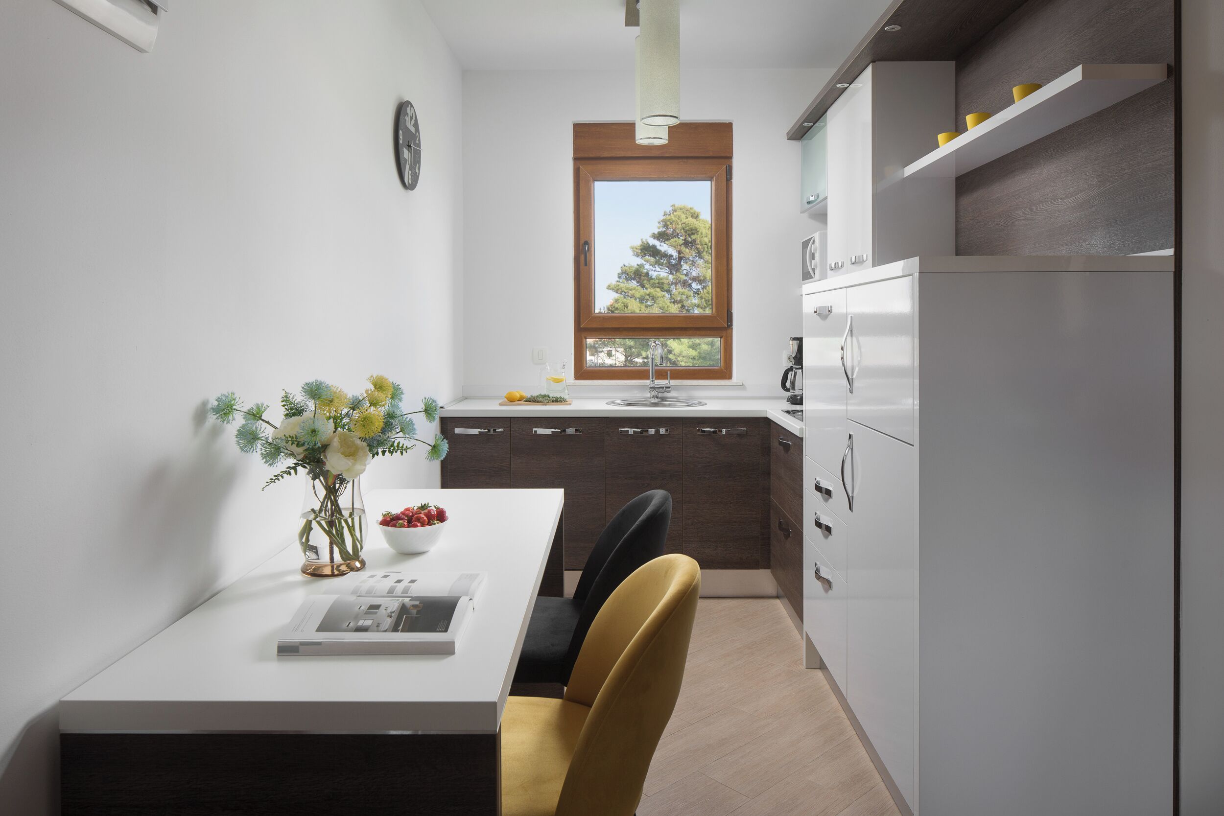 A modern kitchen with a white countertop, a window with a view of trees, and a dining area with a table and chairs.