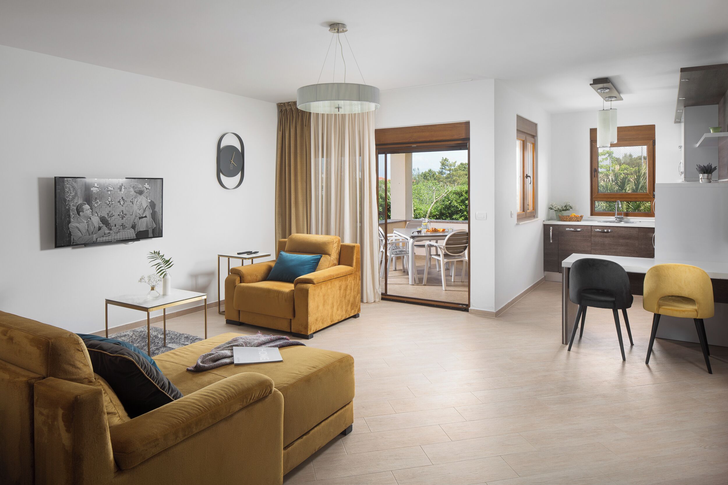 A modern living room with a sofa, a chair, a small table, and a TV. There is a balcony with a dining area and a kitchen visible through the glass doors.