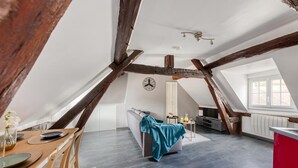 A spacious attic room with exposed wooden beams, a dining area with a wooden table and chairs, a sofa with a teal throw, and a kitchen area with a red countertop. - Drouais Night (Dreux)