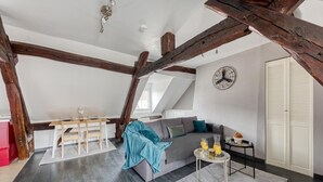 A loft-style room with exposed wooden beams, a gray sofa, a small round table with drinks, and a white radiator. - Drouais Night (Dreux)