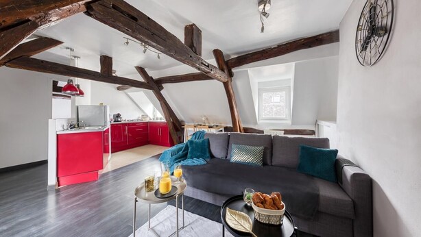 A modern loft apartment with exposed wooden beams, a grey sofa, red kitchen cabinets, and a large window. - Drouais Night (Dreux)