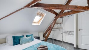 A bedroom with a sloped ceiling, a skylight, a bed with blue and white bedding, a wooden beam structure, and a metal drying rack. - Drouais Night (Dreux)