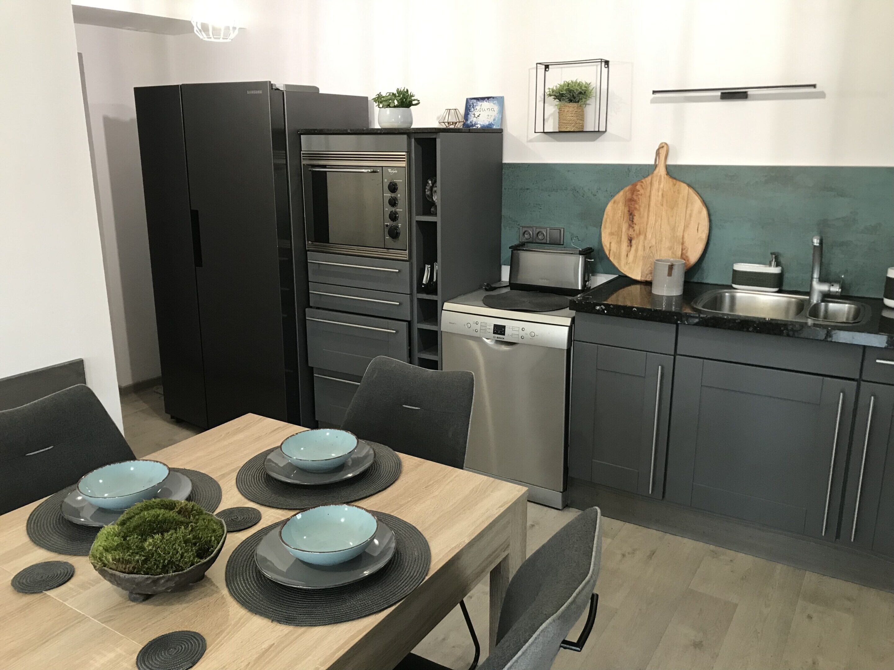 Une cuisine moderne avec un réfrigérateur noir, un four encastré, un lave-vaisselle et une planche à découper en bois posée sur le plan de travail.