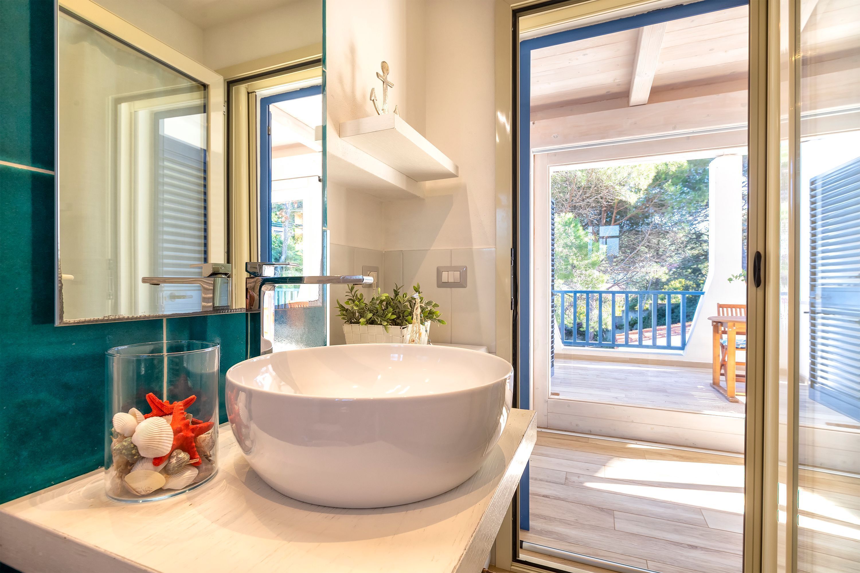 A bathroom with a large white vessel sink, a glass container with decorative items, and a view of the outdoor deck through a glass door.