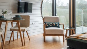 Un interno moderno con soffitto curvo, una TV a schermo piatto montata a parete, una zona pranzo con sedie e tavolo in legno, e un angolo salotto con poltrona e poggiapiedi imbottiti.