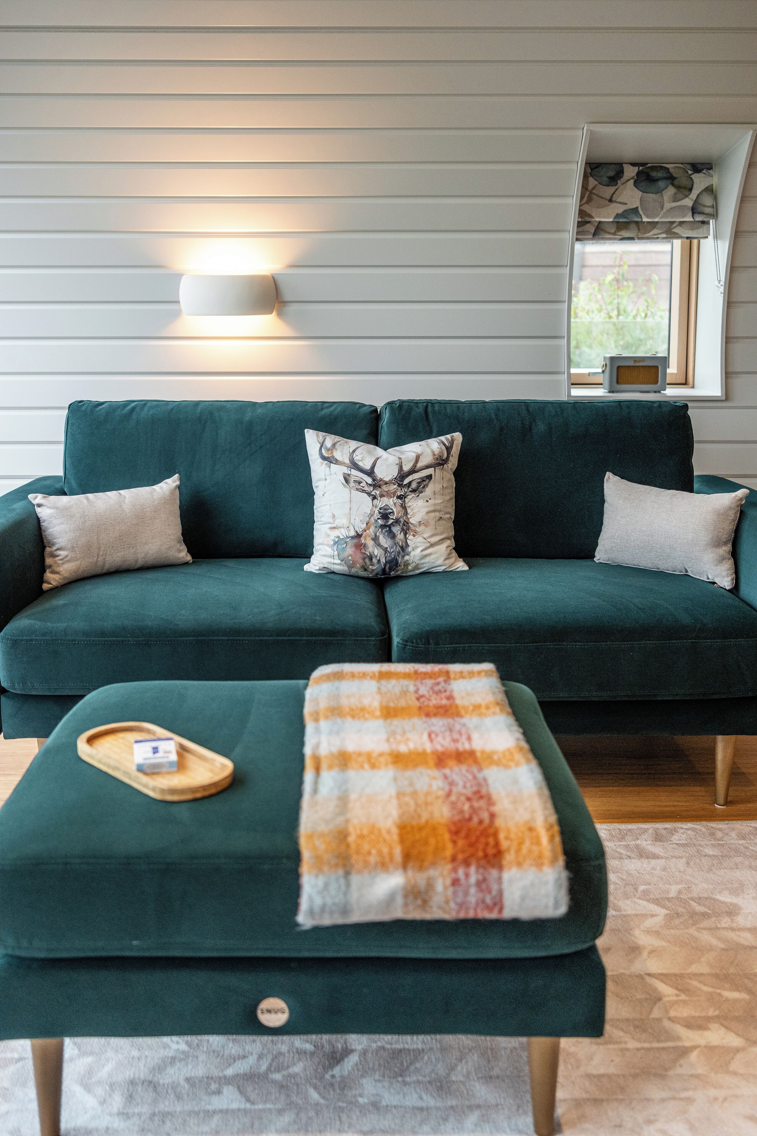 A green sofa with two pillows, one with a deer design, and a matching ottoman with a plaid blanket.