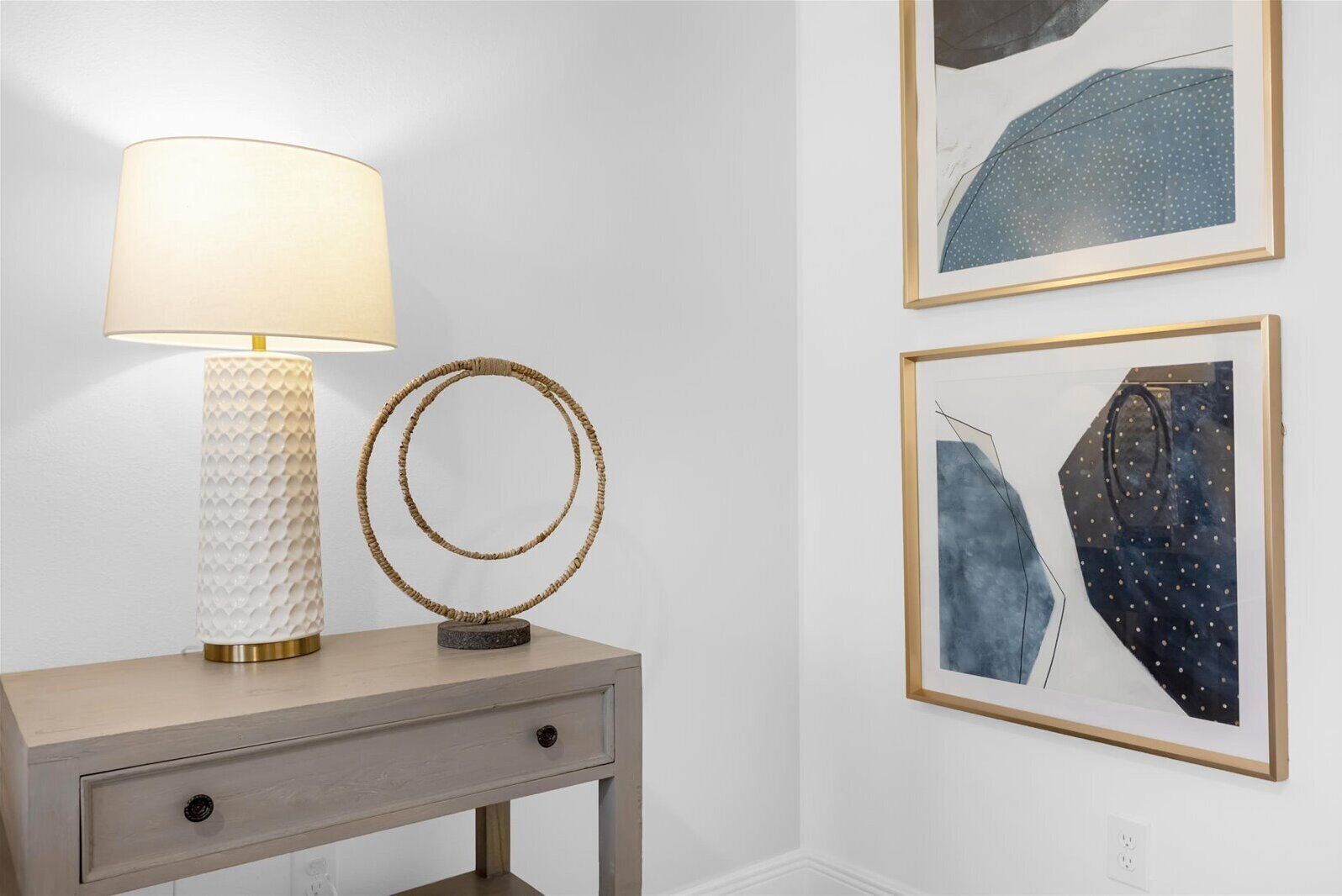 A bedside table with a lamp and a mirror, and two framed artworks on the wall.