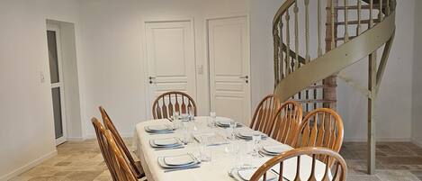 Un coin repas avec une table rectangulaire dressée pour six personnes, entourée de chaises en bois. Un escalier en colimaçon est visible sur la droite.