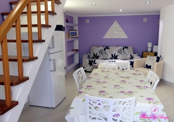 A living room with a sofa, a dining table with chairs, a staircase, and a refrigerator.