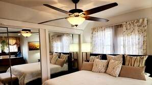 Une chambre d’hôtel avec un grand lit, un ventilateur de plafond, un miroir et une fenêtre agrémentée de rideaux à motifs floraux.