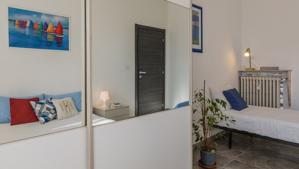 Un dormitorio con cama, mesita de noche, lámpara, planta en maceta, radiador y una puerta.