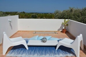 Una terraza en la azotea con una mesa blanca, dos sillas blancas, un tapete azul con estampado y una planta en maceta.