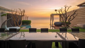 Una mesa con cubierta de vidrio, sillas de mimbre, piscina y vista al mar al atardecer.