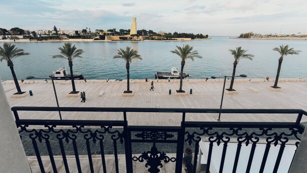 A waterfront promenade with palm trees, a boat dock, and a distant tower. - Verso Oriente  (Brindisi)