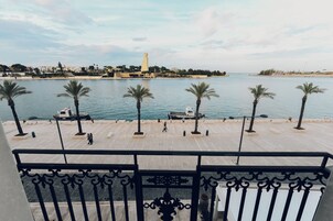 Un paseo marítimo con palmeras, un muelle para barcos y una torre a lo lejos.