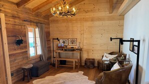Een gezellig blokhutinterieur met houten plafond, een kroonluchter, een bank, een leren fauteuil, een eettafel en een grote bonttapijt.