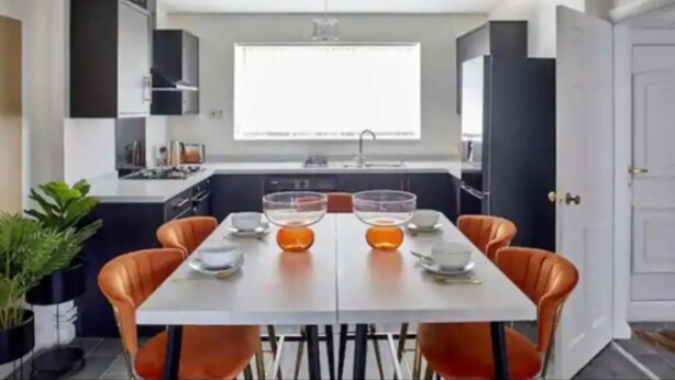A modern kitchen with white cabinets, a large window, and a dining area with orange chairs and a white table set for four.