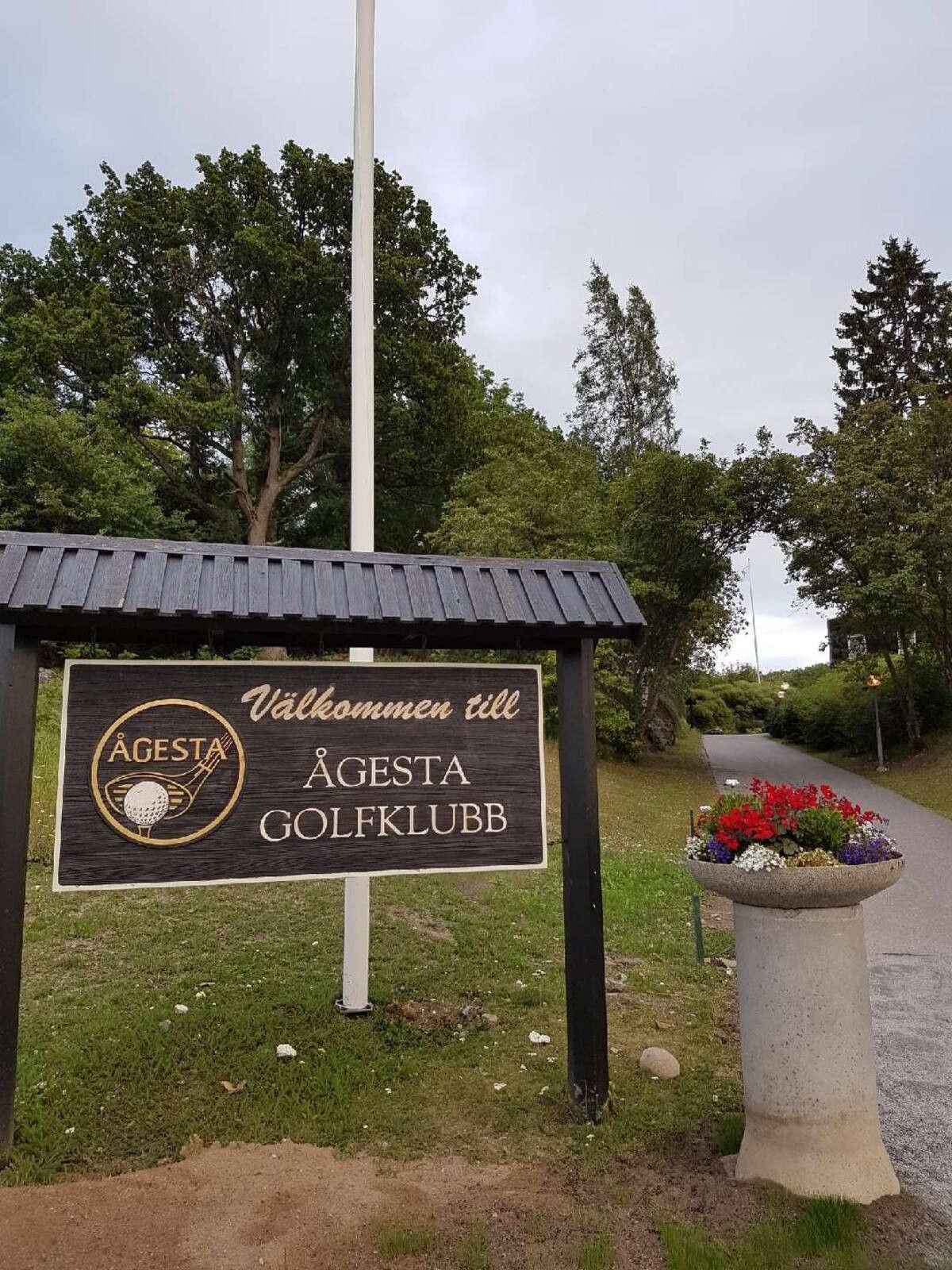 Un senyal de Agesta Golfklubb amb un logotip de pilota de golf, un missatge d'acollida i una jardineria florida al costat d'un camí.