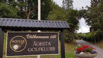 Un senyal de Agesta Golfklubb amb un logotip de pilota de golf, un missatge d'acollida i una jardineria florida al costat d'un camí.
