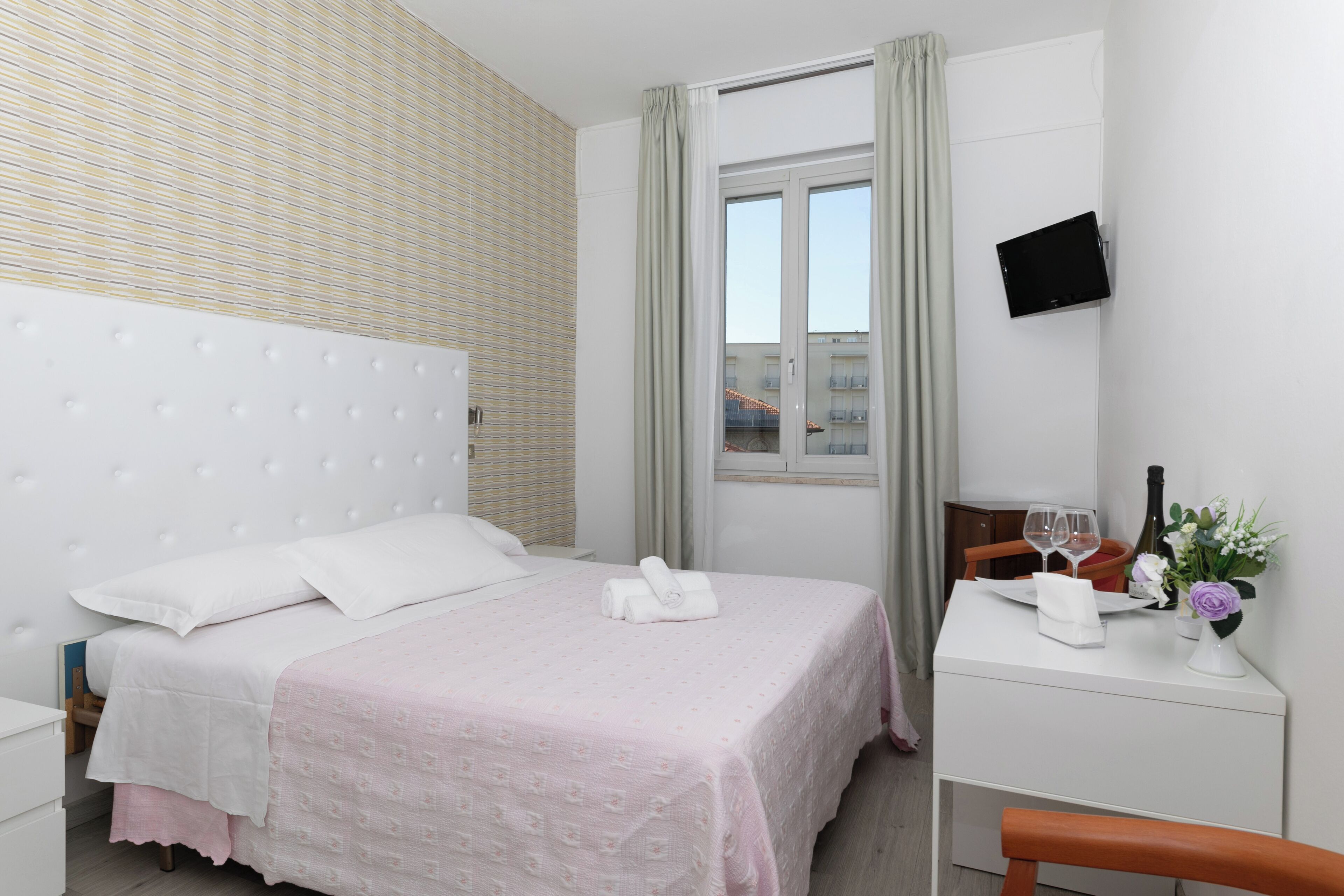 A hotel room with a bed, a bedside table with a bottle, glasses, and a vase of flowers.