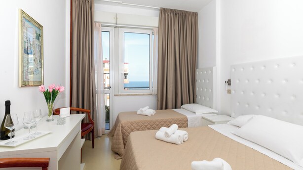 A hotel room with two beds, a desk with a chair, a small table with a bottle and glasses, and a view of the sea through a window. - Hotel La Perla (Rimini)