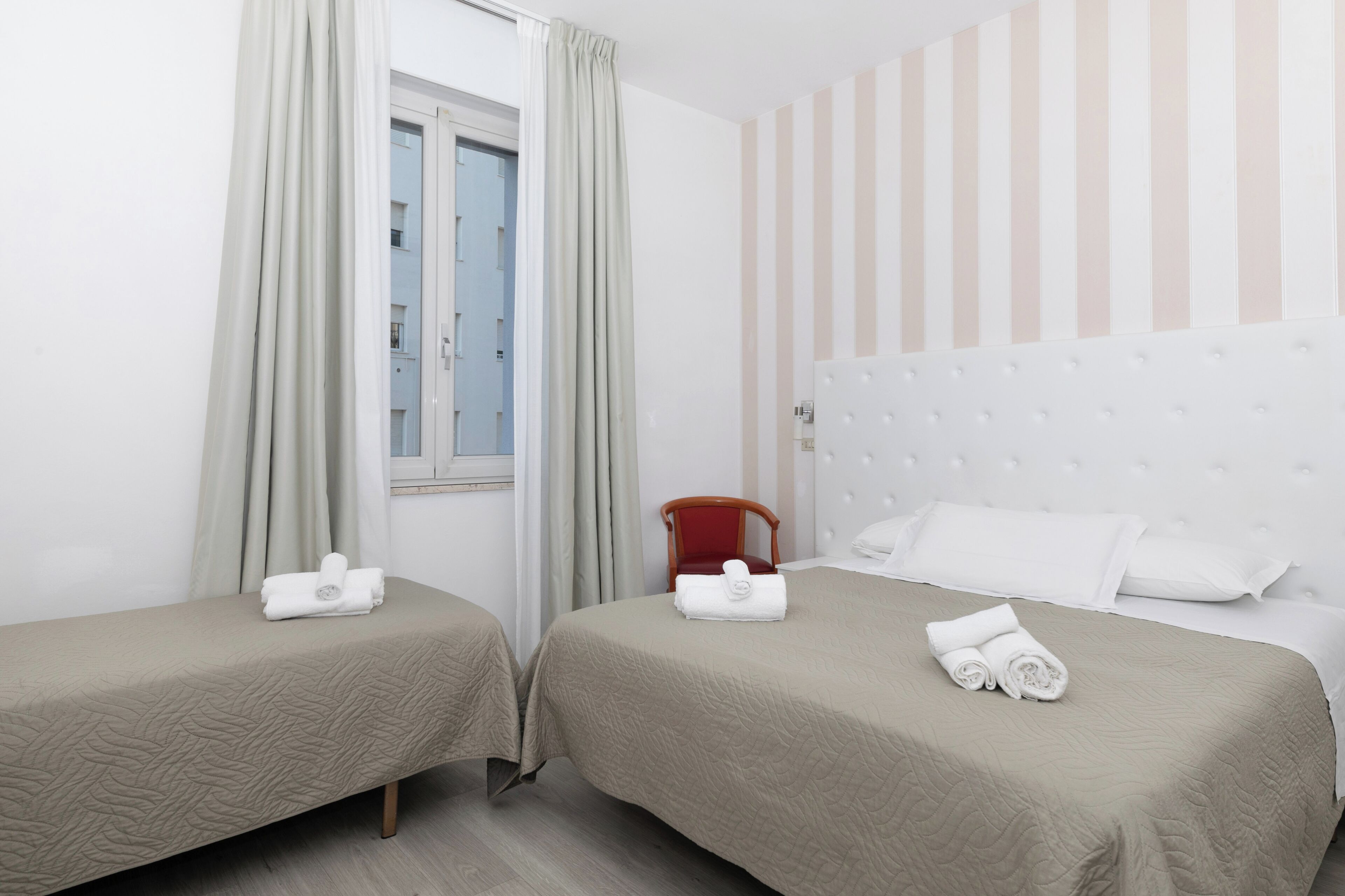 A hotel room with two beds, each with a beige quilted bedspread and a rolled towel. There is a window with curtains, a striped wall, and a red chair.