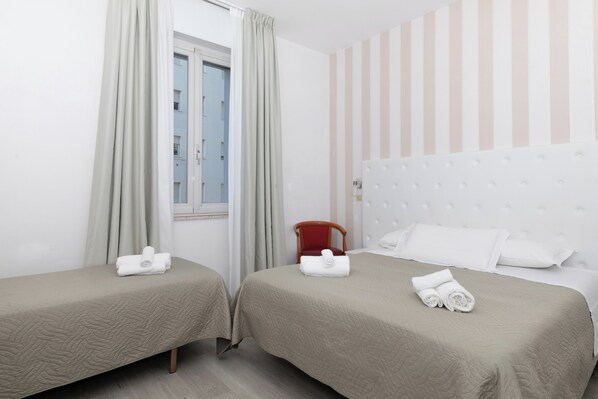 Habitación de hotel con dos camas, cada una con una colcha acolchada color beige y una toalla enrollada. Se ve una ventana con cortinas, una pared a rayas y una silla roja.