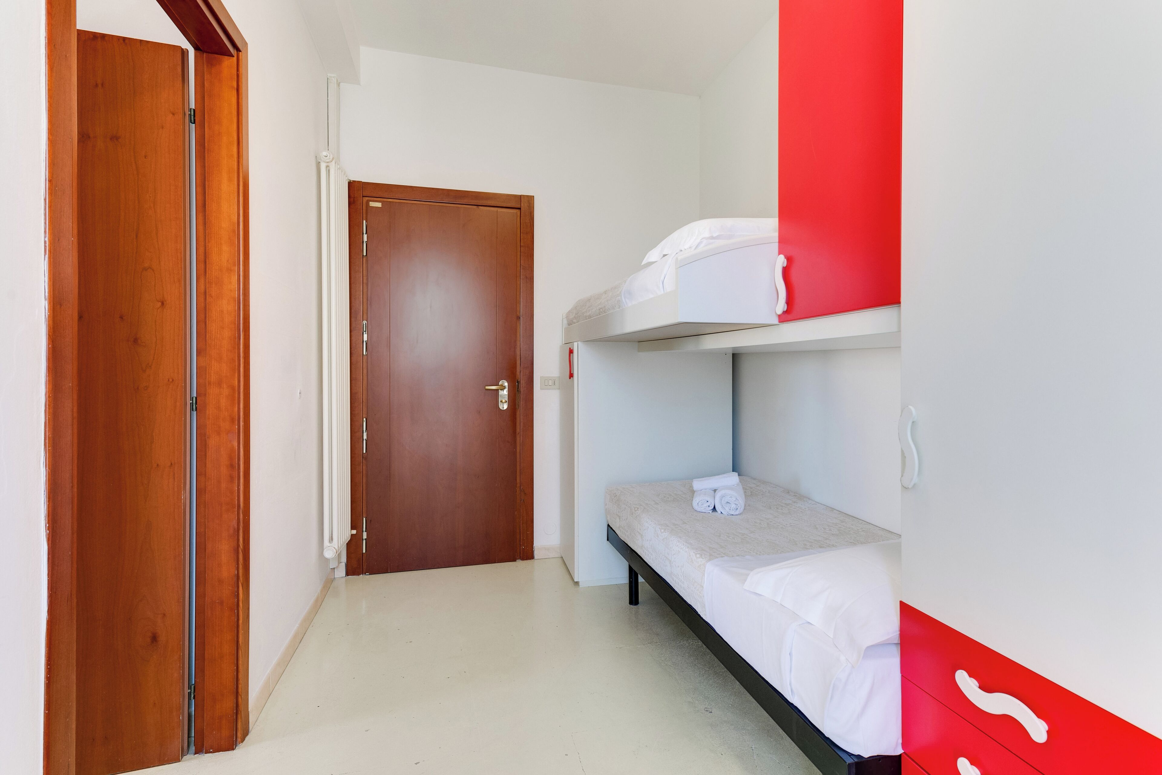 A small, narrow room with a wooden door, a bunk bed with white bedding, and a red accent wall.