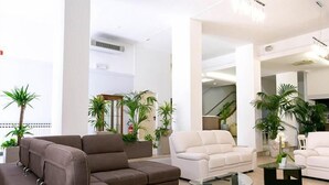 A modern living room with a sectional sofa, a white armchair, and a glass coffee table. There are several potted plants and a staircase in the background. - Hotel La Perla (Rimini)