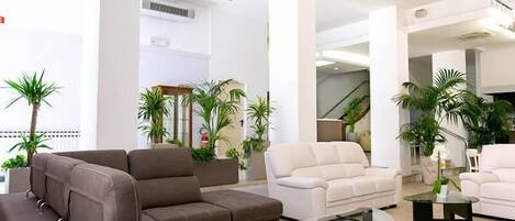 A modern living room with a sectional sofa, a white armchair, and a glass coffee table. There are several potted plants and a staircase in the background.
