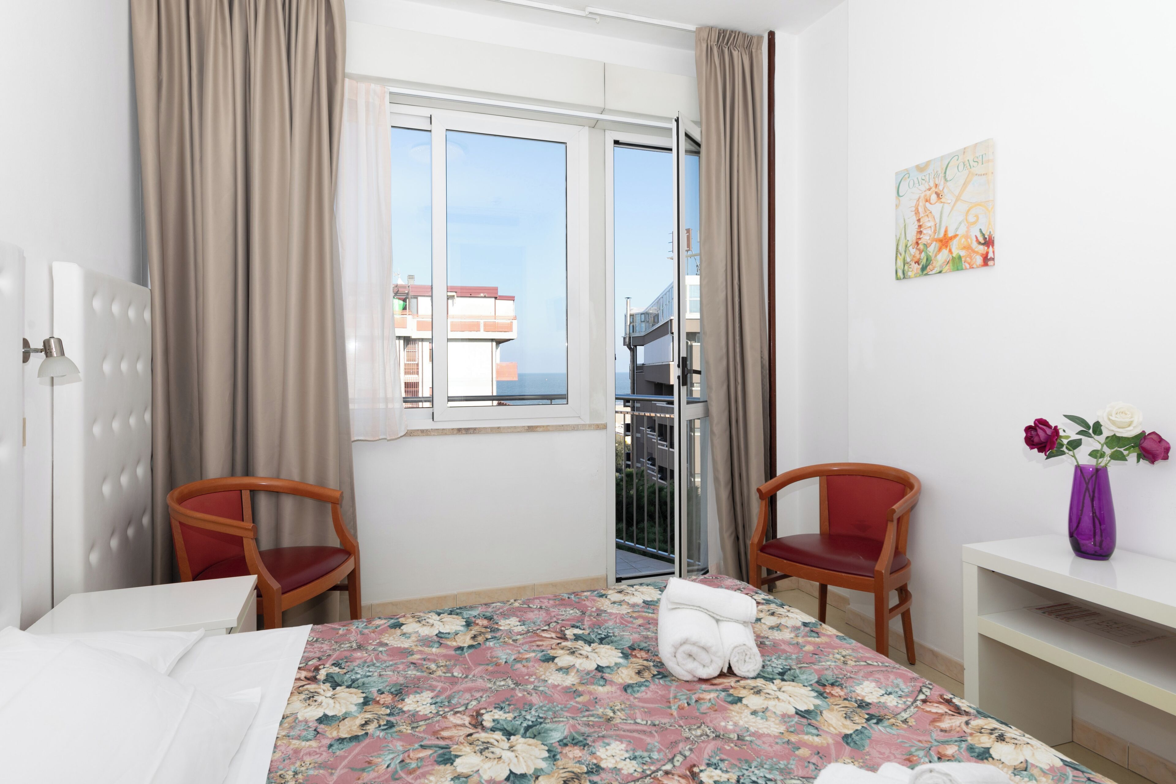 Habitació d'hotel amb un llit, dues cadires, escriptori, un vas amb flors i finestra amb vista a edificis.