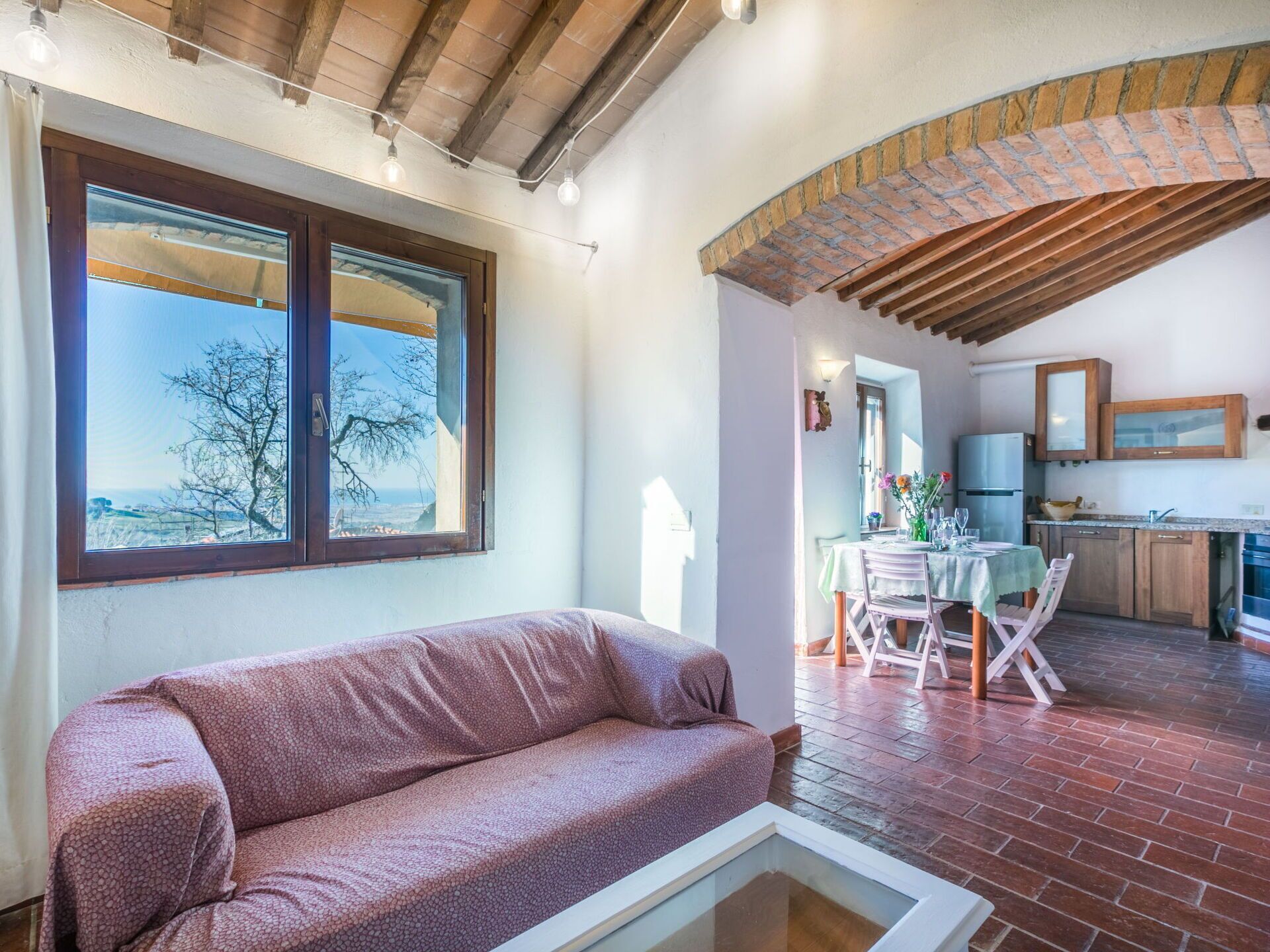 Un accogliente soggiorno con divano, una zona pranzo con tavolo e sedie, una cucina con pensili e frigorifero, e un'ampia finestra con vista sugli alberi.