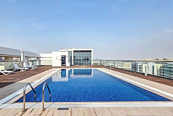 A rooftop swimming pool with a clear view of the city skyline, lounge chairs, and a glass-enclosed building.