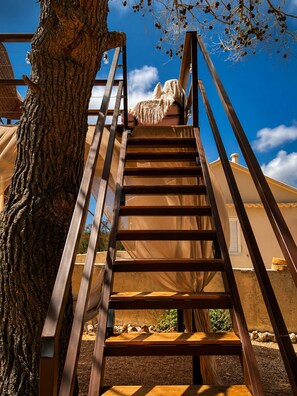 Una escalera de madera con barandillas metálicas que conduce a un edificio de fachada color beige y un árbol con ramas desnudas.