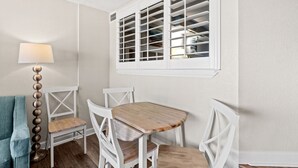 A dining area with a wooden table and white chairs, a lamp, and a window with blinds. - Oceanfront Condo with Resort Pools (North Myrtle Beach)