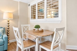 A dining area with a round wooden table, white chairs, a small potted plant, and a window with blinds.