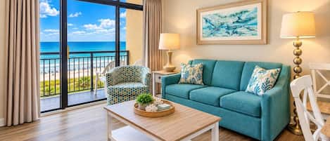 A room with a view of the sea, a teal sofa, a patterned armchair, a wooden coffee table, and two lamps.