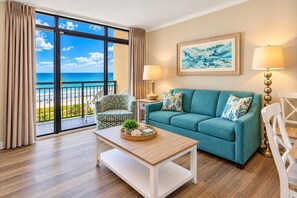 A room with a view of the sea, a teal sofa, a patterned armchair, a wooden coffee table, and two lamps. - Oceanfront Condo with Resort Pools (North Myrtle Beach)