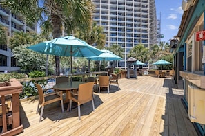 A wooden deck with outdoor seating, palm trees, and a building in the background.