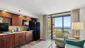 A modern kitchen with wooden cabinets, a black refrigerator, and a microwave. There is a dining table and a chair with a colorful pattern. A large window offers a view of the ocean. - Oceanfront Condo with Resort Pools (North Myrtle Beach)