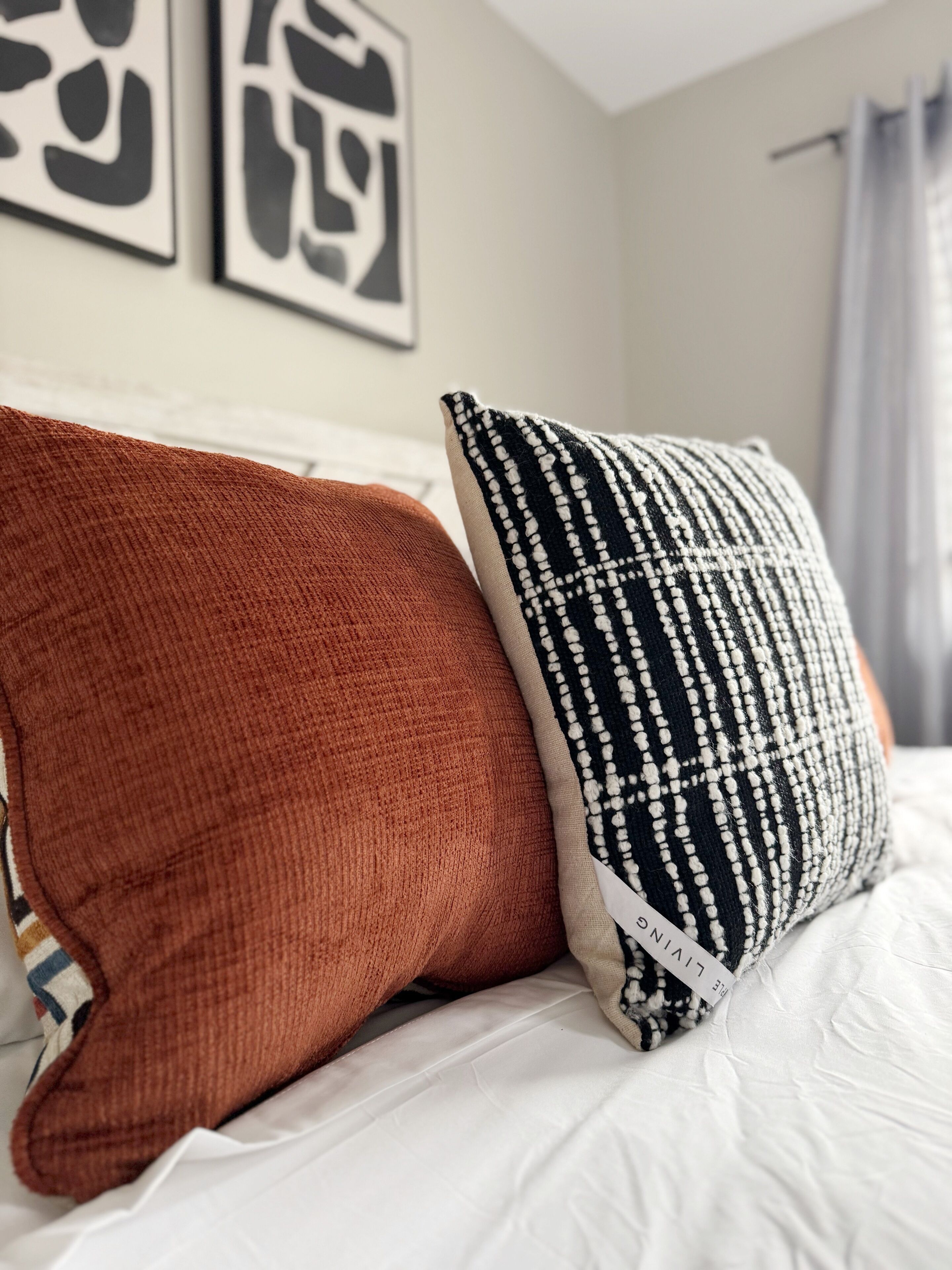 Una cama con dos almohadas, una marrón y otra con estampado blanco y negro, en una habitación con una pared blanca y un cuadro enmarcado sobre la cama.