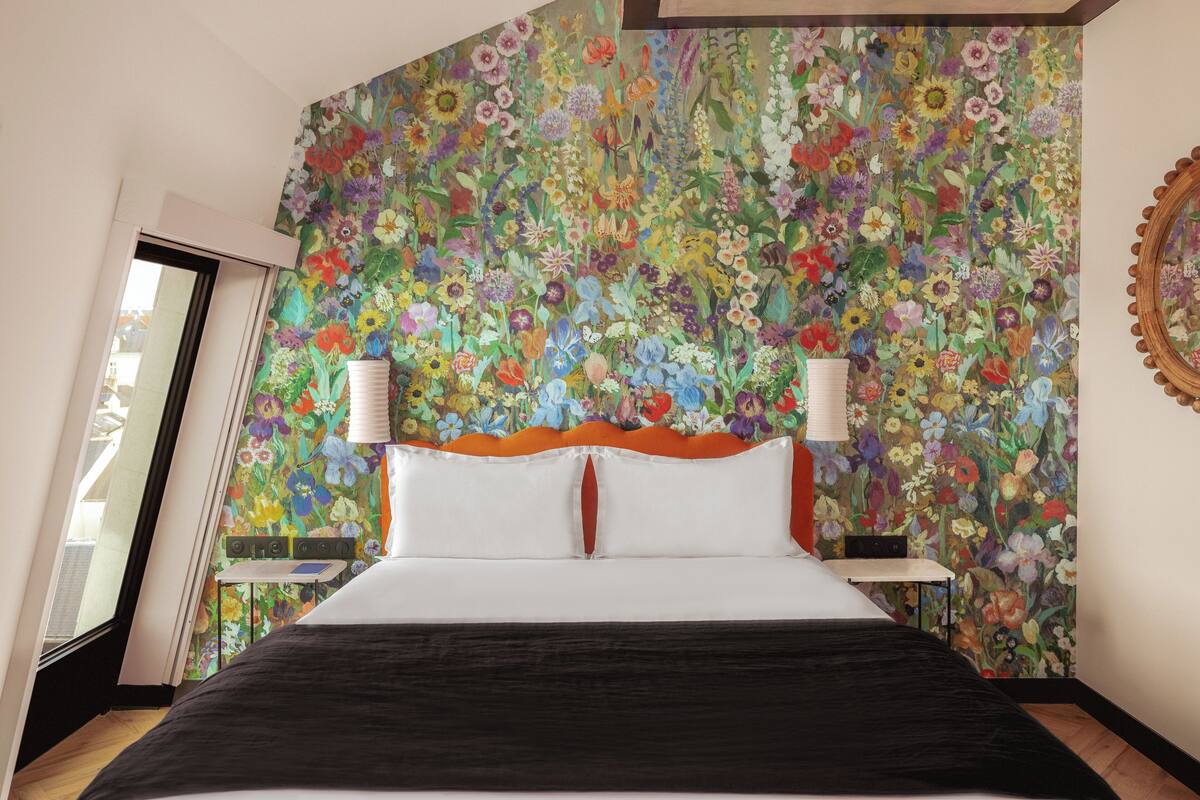 A bedroom with a floral wallpaper, a wooden headboard, two bedside lamps, and a window with a view.
