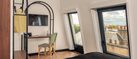 A modern bedroom with a bed, a desk, and a chair. There is a large window providing a view of the cityscape.