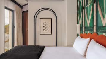 A hotel room with a bed, a framed artwork with the text "cha cha cha", a geometric patterned wall, and a window with curtains.