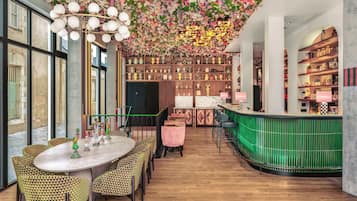 Amplia habitación con techo alto decorado con motivos florales, una zona de bar con mostrador verde y un espacio de estar con sillas estampadas y una mesa redonda.