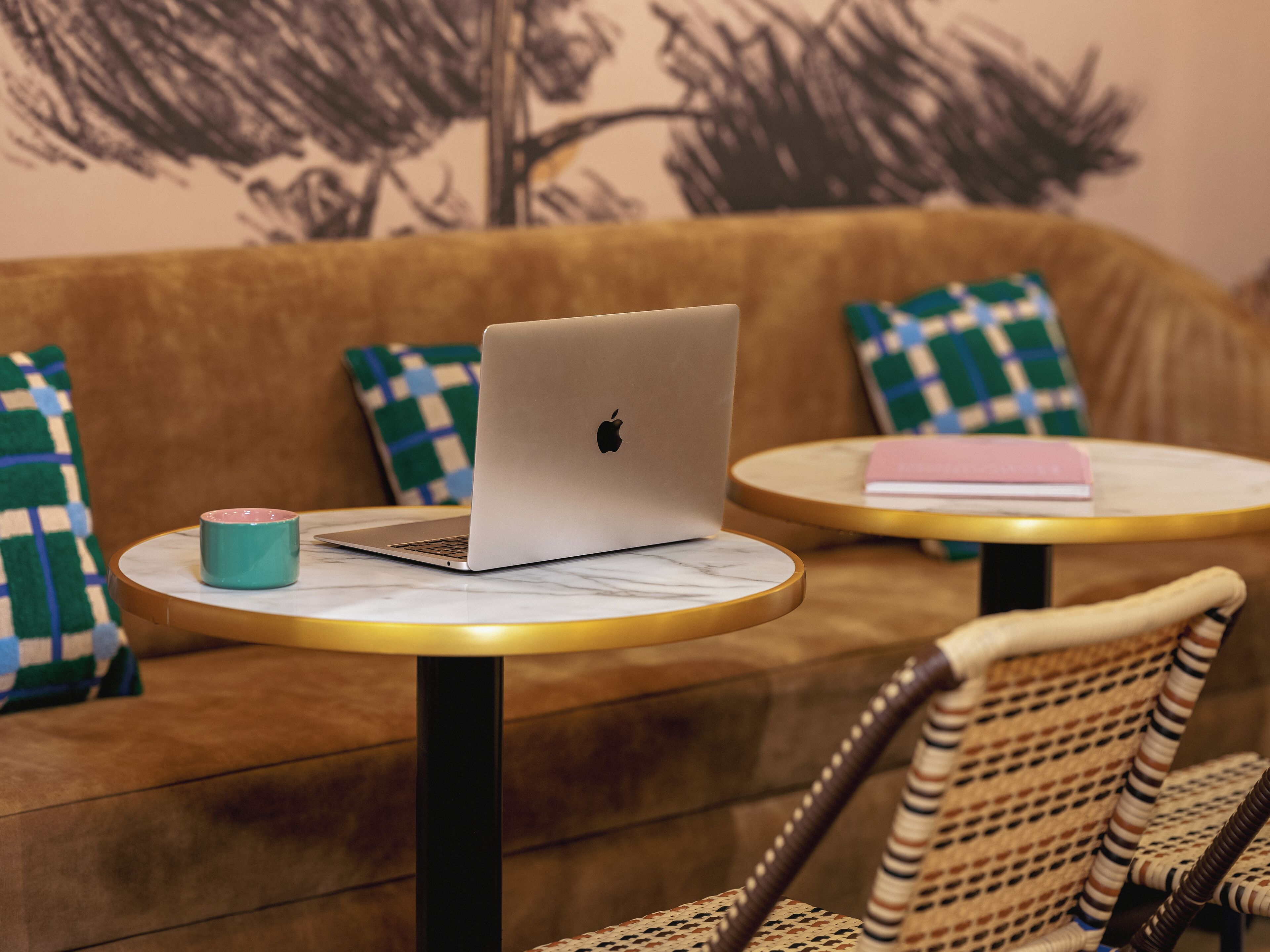 A laptop on a round table with a checkered pillow, a book, and a mug.