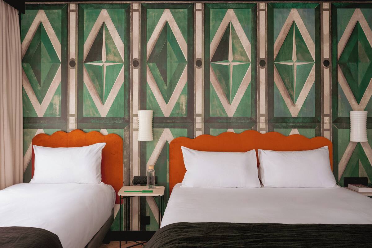 A hotel room with two beds, each with an orange headboard and white bedding. The walls feature a geometric pattern with green and beige tones.