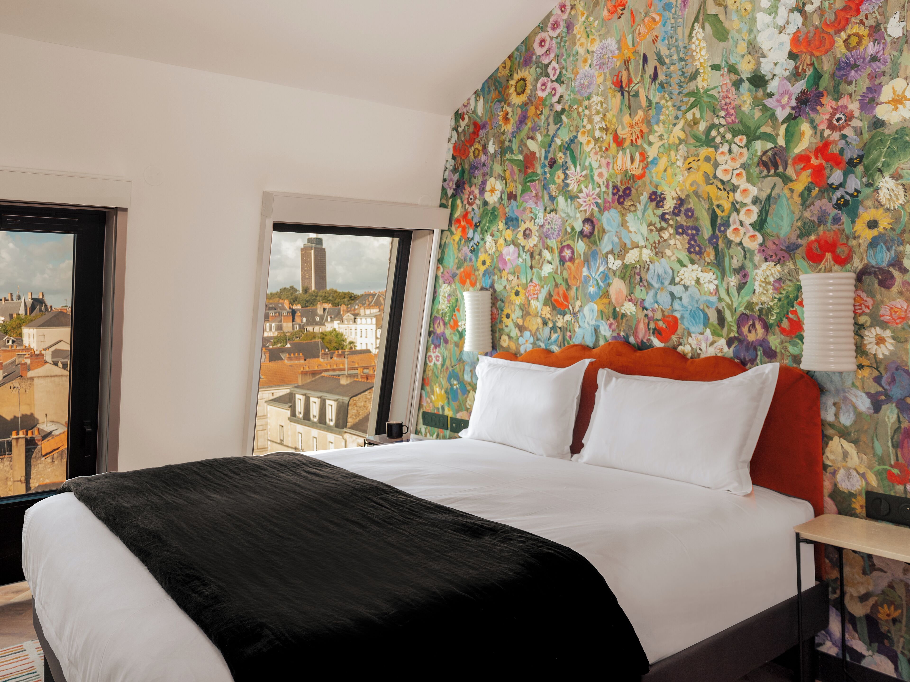 A hotel room with a large bed, a view of the city through a window, and a floral wallpaper on the wall.